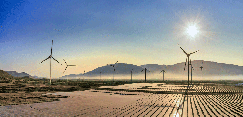 Image of an industrial solar farm outside with windmills, mountains, and the setting sun in the background.